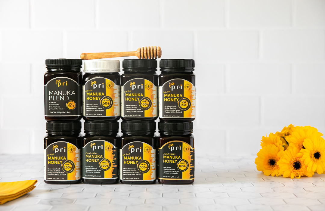 Assorted jars of Manuka honey stacked with a honey dipper and yellow flowers on a white background.