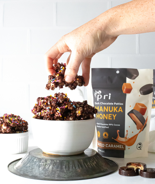 Hand holding chocolate Manuka honey cluster with sprinkles above bowl, Manuka honey product pouch in background