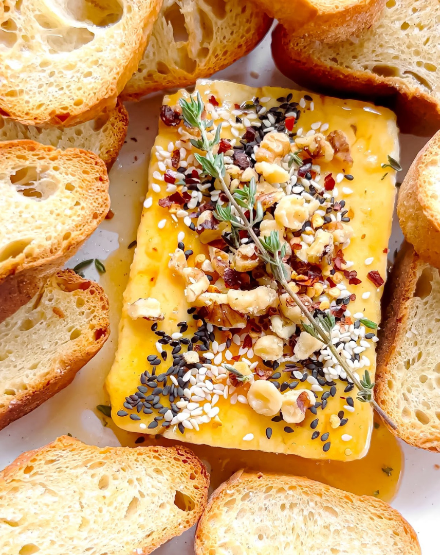 Honey-drizzled cheese block topped with nuts, sesame seeds, herbs, surrounded by toasted baguette slices
