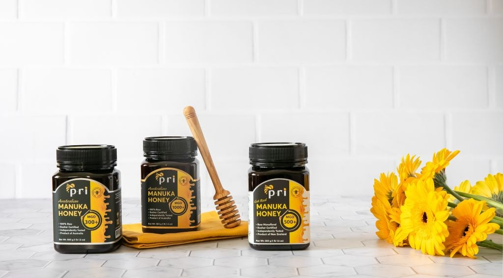 Three jars of Manuka honey with different MGO ratings, honey dipper, and yellow flowers on a white counter.