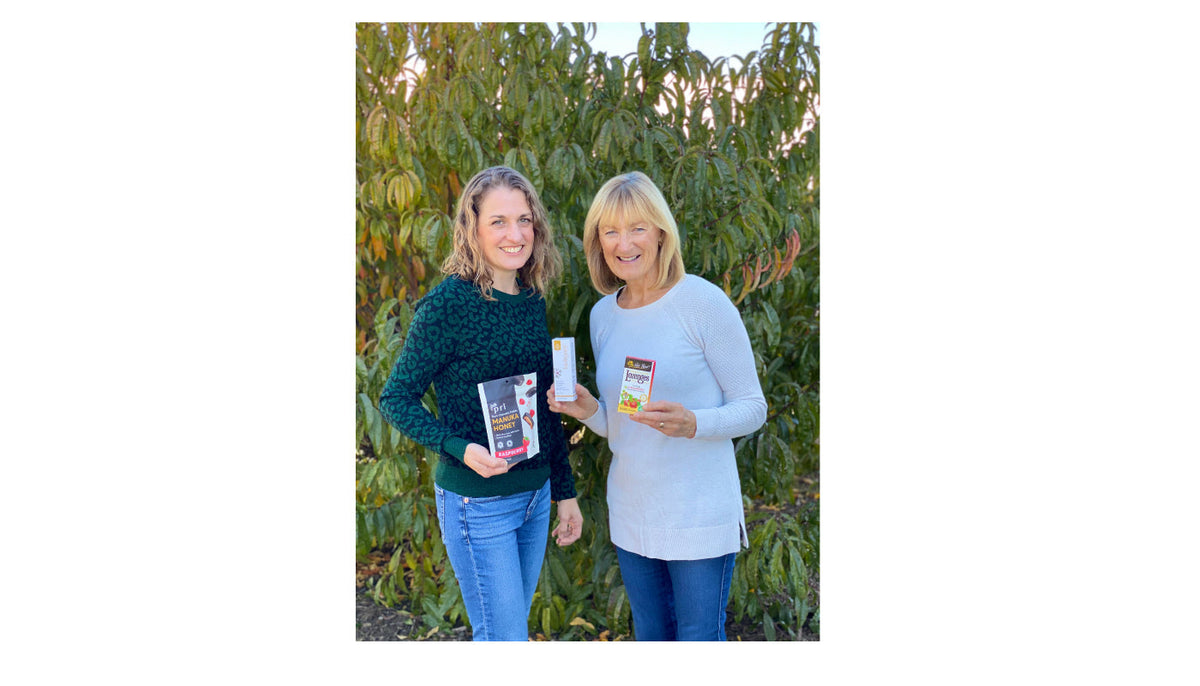 Two women outdoors holding Manuka honey and wellness products in front of green foliage
