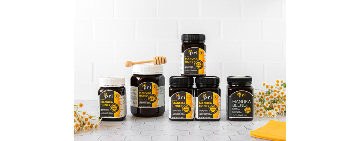Assorted jars of Manuka honey and Manuka blend on a counter with flowers and honey dipper