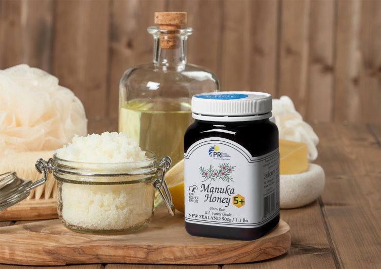 Jar of Manuka honey with sugar scrub, oil bottle, and spa accessories on wooden tray