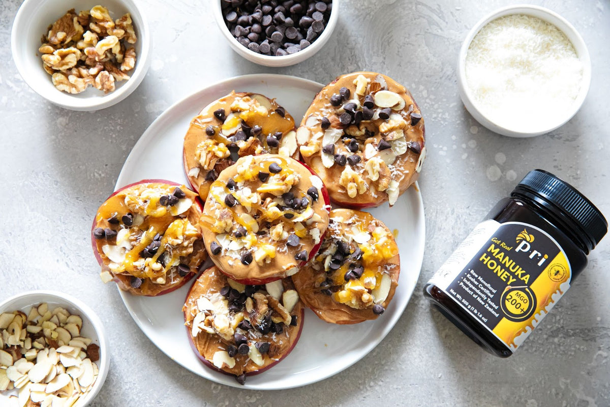 Apple slices with peanut butter, Manuka honey, nuts, chocolate chips, and coconut on a plate, with a jar of Manuka honey nearby.
