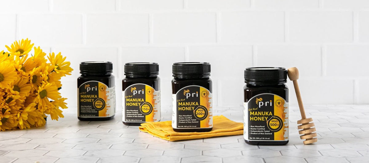 Four jars of Manuka honey with yellow flowers and honey dipper on a white kitchen counter